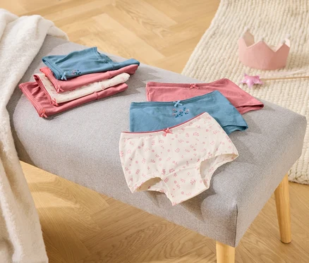 Sur une banquette grise sont posés des slips pour filles pliés, roses, blancs et bleus, à côté d'une couronne jouet et d'une baguette.
