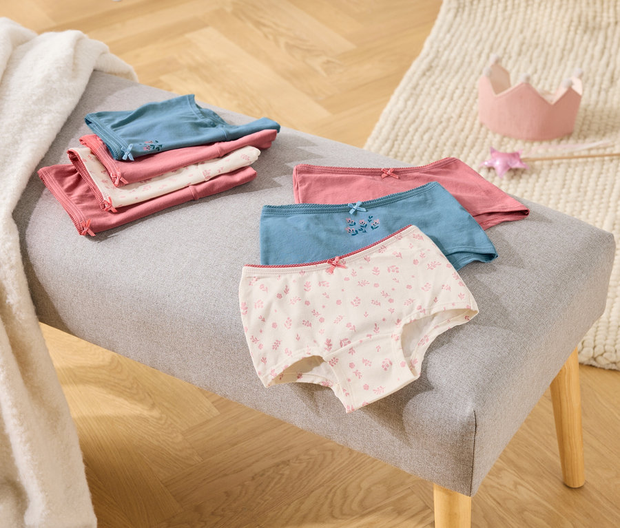 Sur une banquette grise sont posés des slips pour filles pliés, roses, blancs et bleus, à côté d'une couronne jouet et d'une baguette.