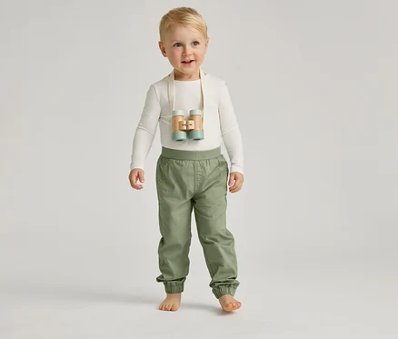 Un garçon aux pieds nus avec des yeux bleus et des cheveux blonds se tient debout avec un pantalon clair et un haut clair sur un fond gris.