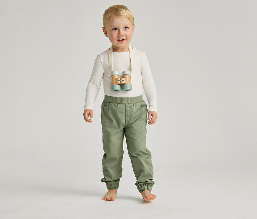 Un garçon aux pieds nus avec des yeux bleus et des cheveux blonds se tient debout avec un pantalon clair et un haut clair sur un fond gris.