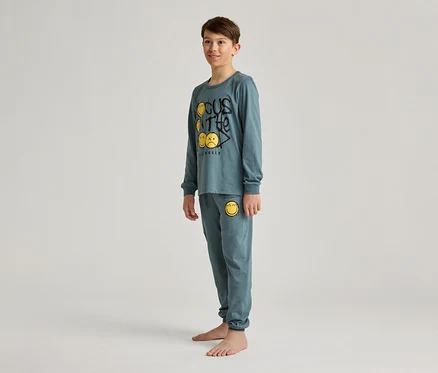 Un garçon se tient pieds nus dans un pyjama bleu-vert avec des smileys jaunes et l'inscription « Focus on the real world ».