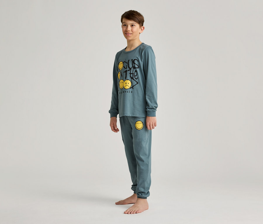 Un garçon se tient pieds nus dans un pyjama bleu-vert avec des smileys jaunes et l'inscription « Focus on the real world ».