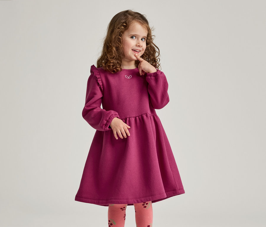 Une fillette aux cheveux bouclés porte une robe bordeaux à volants et des collants à motifs, touchant pensivement son doigt.