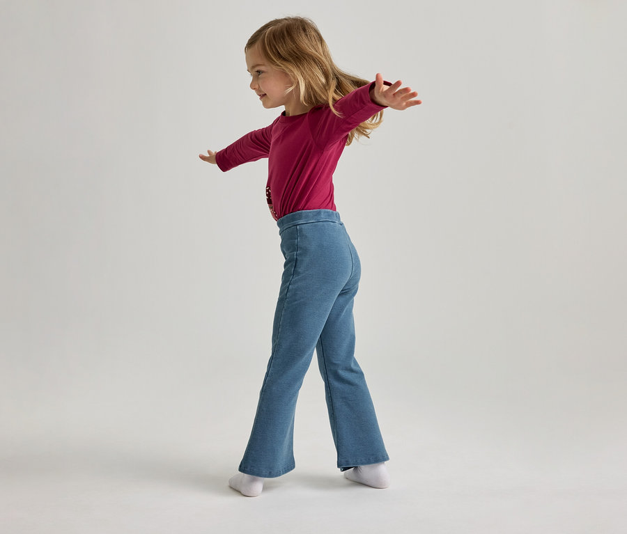 Fillette portant un haut rose à manches longues et un pantalon bleu évasé, les bras tendus, vue latérale.