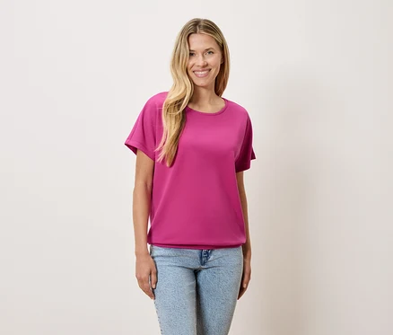 Une femme aux cheveux blonds porte un t-shirt rose et un jean bleu.