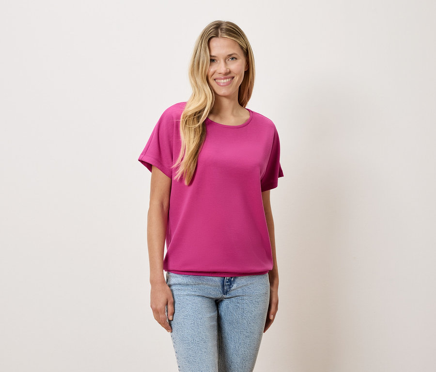 Une femme aux cheveux blonds porte un t-shirt rose et un jean bleu.