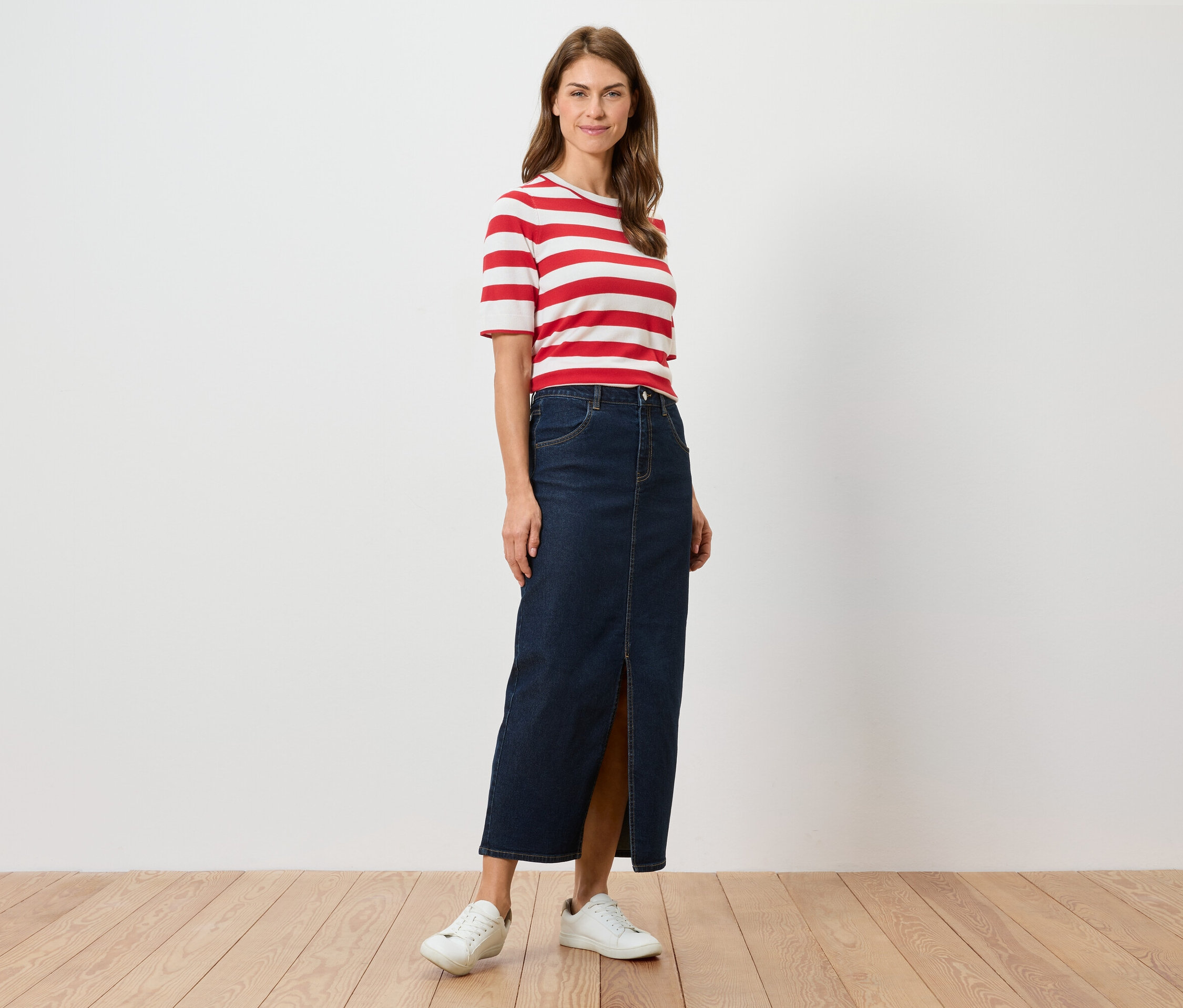 Une femme debout porte un t-shirt rayé rouge et blanc, une longue jupe en jean fendue et des baskets blanches.