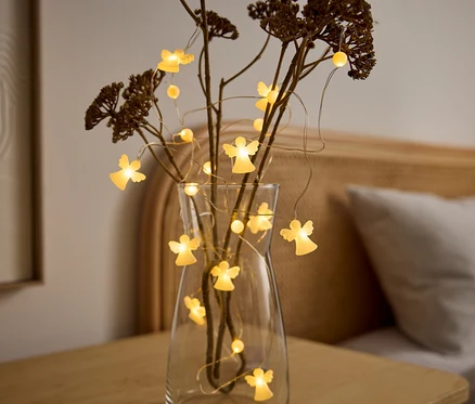Un vase en verre transparent contient des branches séchées et une guirlande lumineuse avec des figurines d'anges éclairées. Le vase est posé sur une table en bois, avec un lit en arrière-plan.