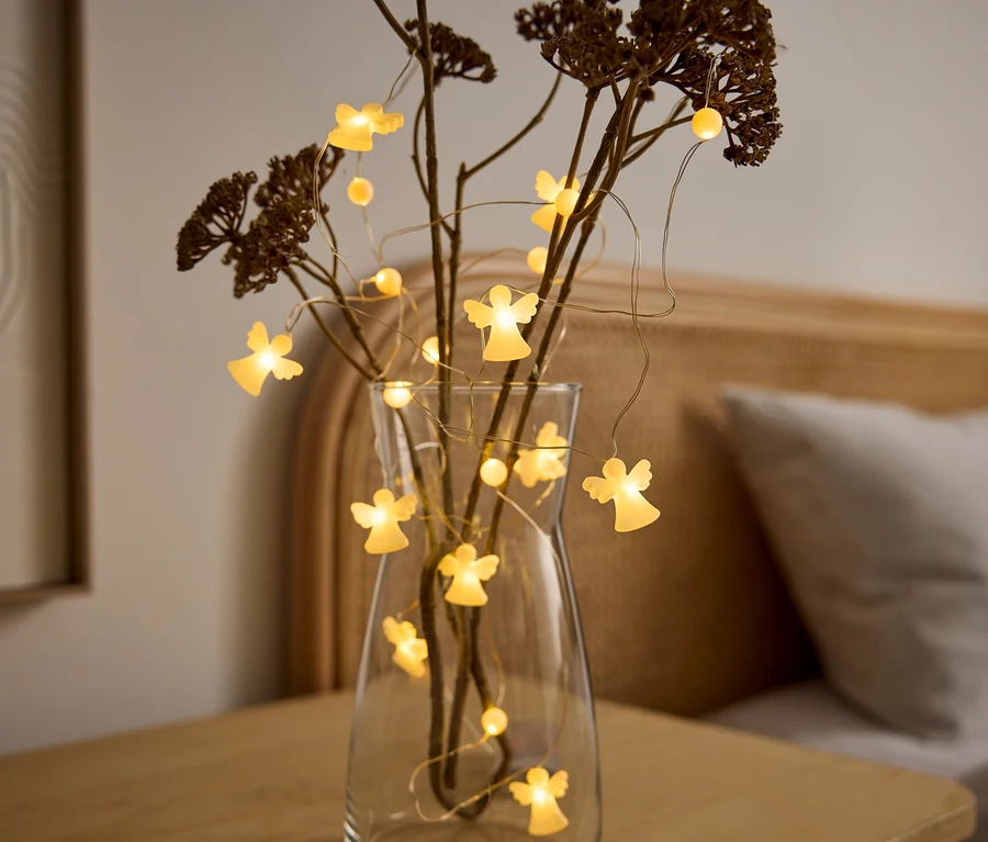 Un vase en verre transparent contient des branches séchées et une guirlande lumineuse avec des figurines d'anges éclairées. Le vase est posé sur une table en bois, avec un lit en arrière-plan.