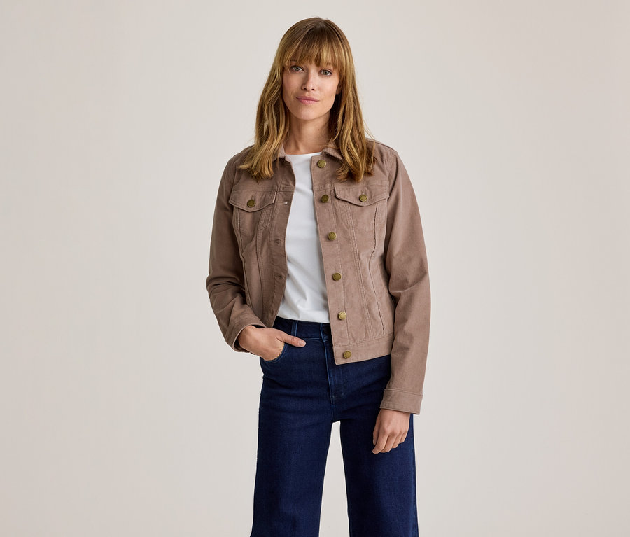 Femme portant une veste en velours côtelé marron avec des boutons en laiton, un t-shirt blanc et un jean large bleu foncé, debout la main dans la poche.