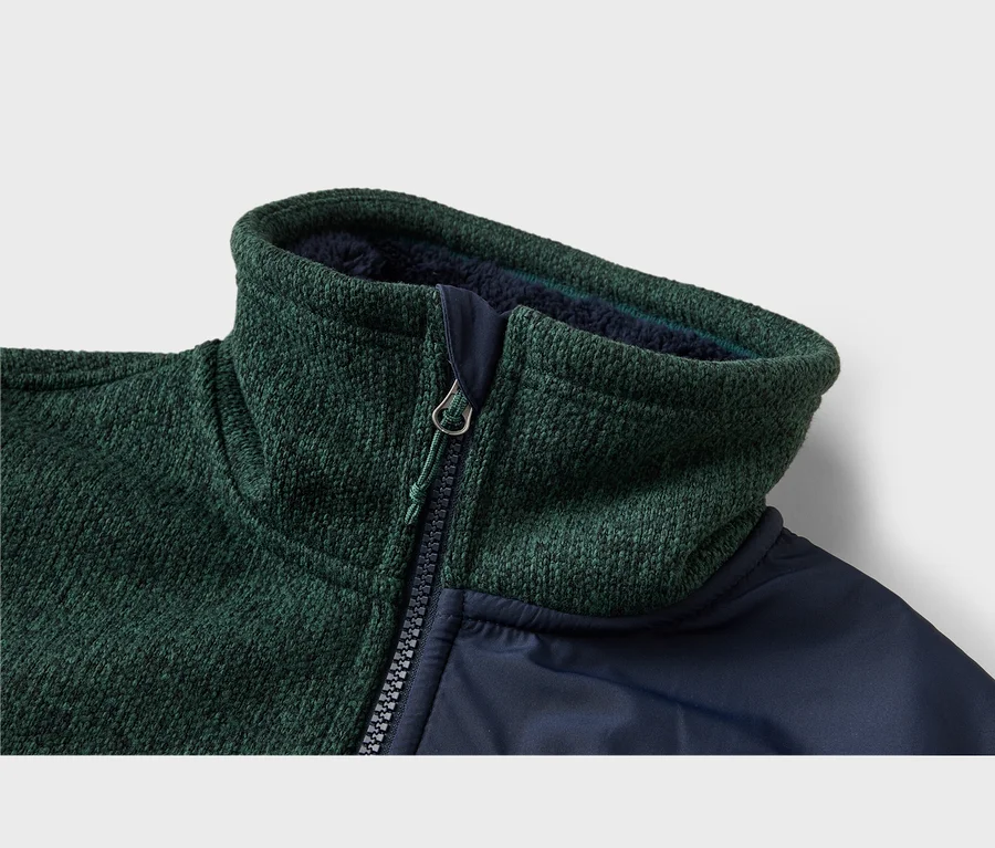 Gros plan d'une veste texturée vert foncé avec un tissu lisse bleu foncé. Col montant avec doublure bleu foncé et fermeture éclair noire avec tirette argentée et cordon vert.