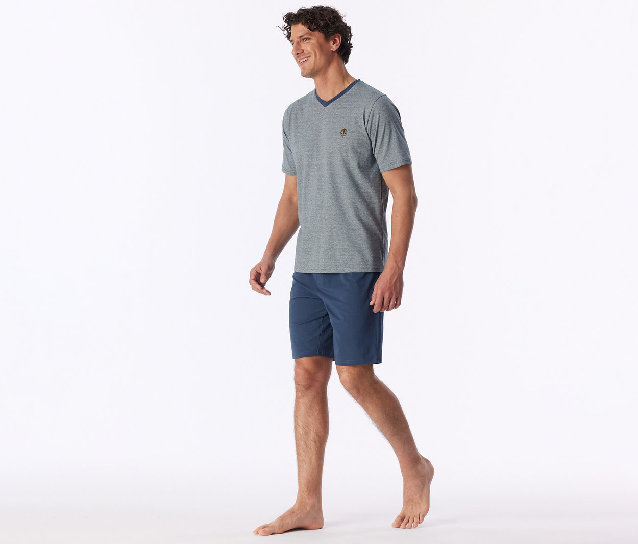 Un homme souriant aux cheveux bruns bouclés marche pieds nus, vêtu d'un t-shirt gris à col en V avec un petit emblème et d'un short bleu marine.