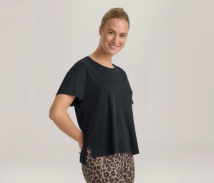 Femme souriante aux cheveux attachés, portant un t-shirt noir et un legging imprimé léopard, les mains derrière le dos.
