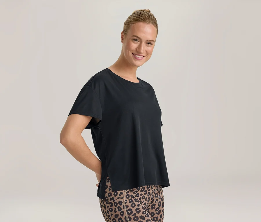 Femme souriante aux cheveux attachés, portant un t-shirt noir et un legging imprimé léopard, les mains derrière le dos.