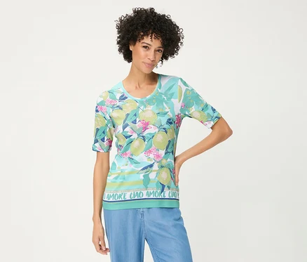 Femme aux cheveux bouclés posant, vêtue d'un T-shirt Olsen à fond bleu clair, motifs de fruits d'agrumes verts, fleurs roses et texte « AMORE CIAO » à l'ourlet, associé à un pantalon bleu clair.