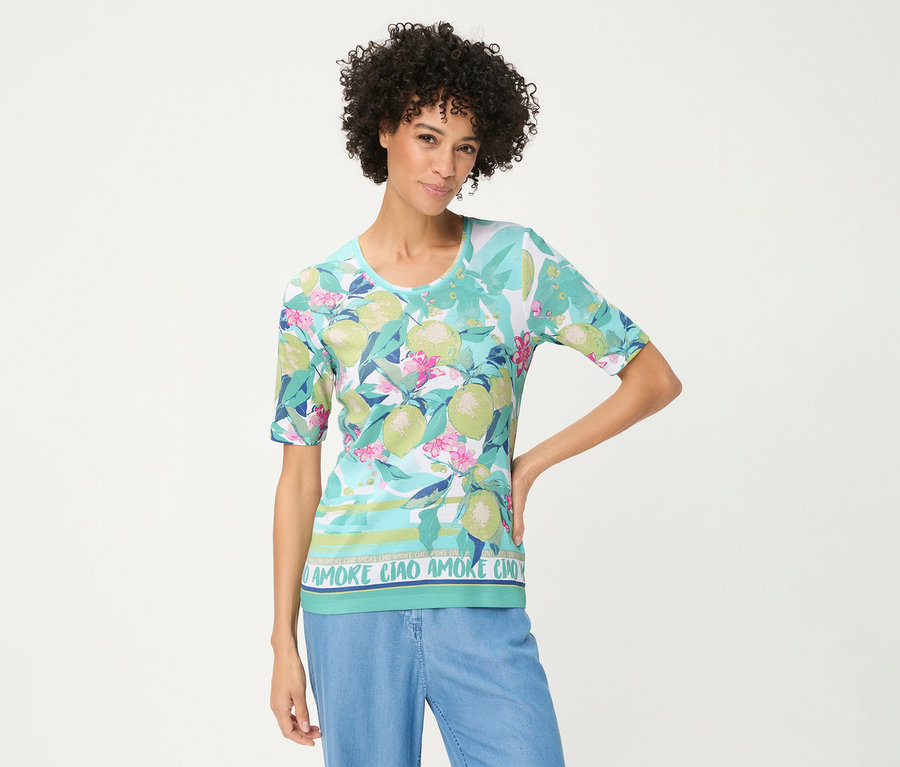 Femme aux cheveux bouclés posant, vêtue d'un T-shirt Olsen à fond bleu clair, motifs de fruits d'agrumes verts, fleurs roses et texte « AMORE CIAO » à l'ourlet, associé à un pantalon bleu clair.