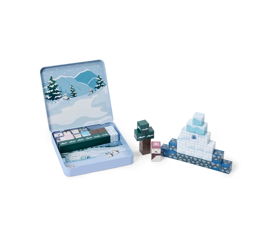 Une boîte en étain bleu clair ouverte avec des illustrations de scène hivernale. À l'intérieur se trouvent des rangées de petits blocs de construction. À droite, des blocs vert foncé forment un arbre, deux blocs à têtes d'animaux sont seuls, et des blocs blancs et bleus forment un igloo sur une base bleu foncé.