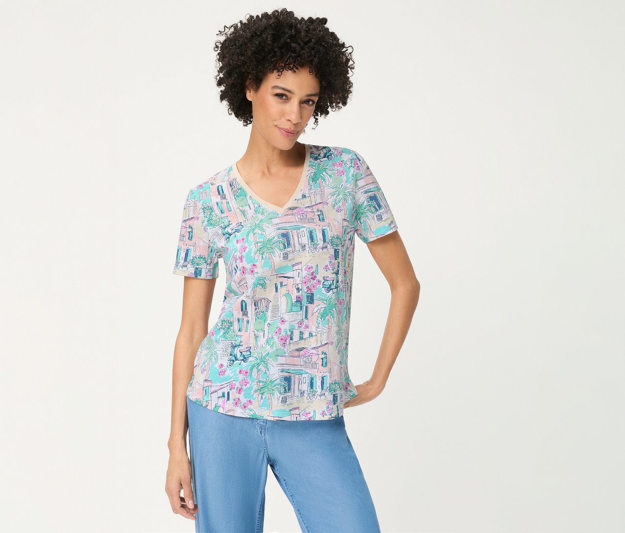 Femme aux cheveux bouclés portant un T-shirt Olsen à imprimé pastel de bâtiments, palmiers et fleurs, avec un pantalon bleu clair, souriant la main sur la hanche.