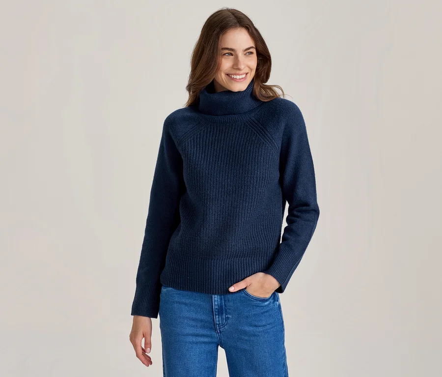 Femme souriante aux longs cheveux bruns, portant un pull col roulé côtelé bleu marine et un jean bleu, une main dans la poche.