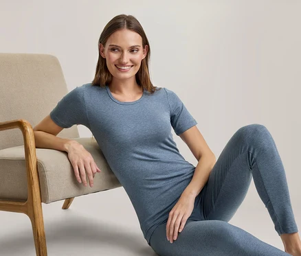 Une femme souriante est assise par terre, appuyée contre un fauteuil beige, vêtue d'un t-shirt bleu et d'un legging assorti.