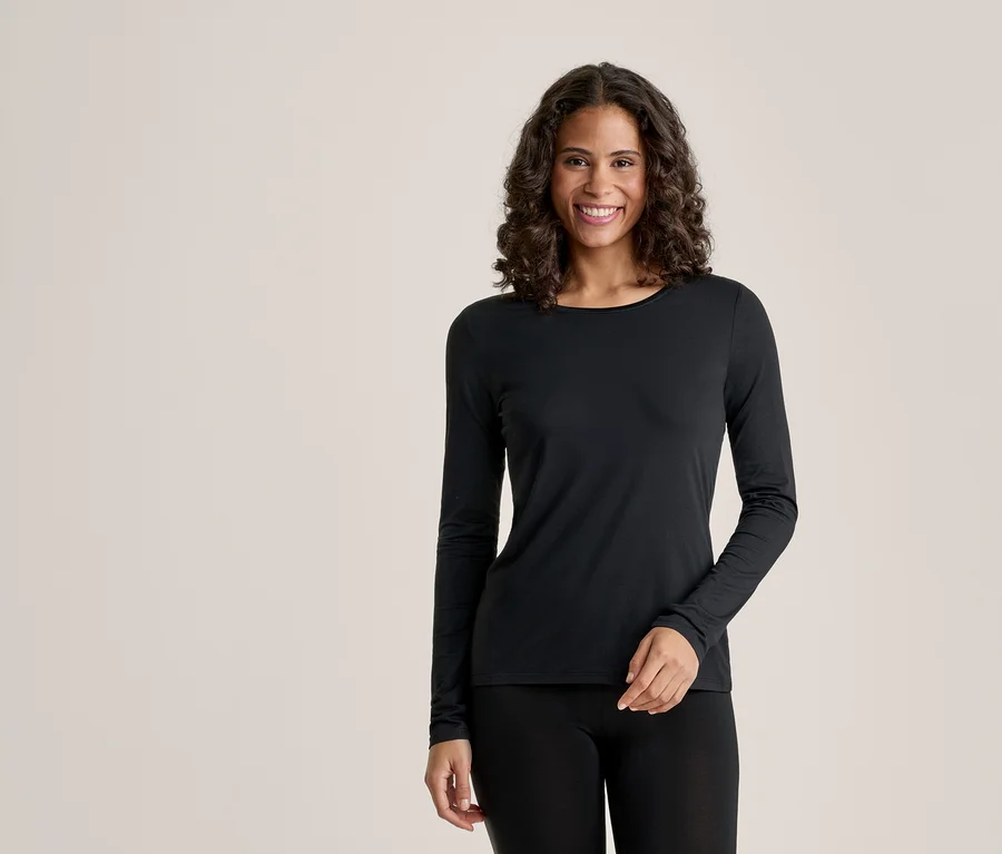 Une femme souriante aux cheveux bouclés foncés porte un haut noir à manches longues et un legging noir, debout devant un fond clair.