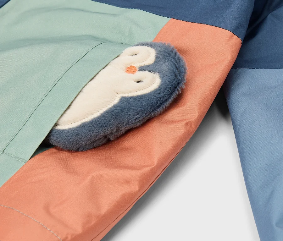 Gros plan d'un vêtement color block bleu foncé, vert clair et pêche, avec un pingouin en peluche bleu et blanc au bec orange partiellement glissé dans une couture.