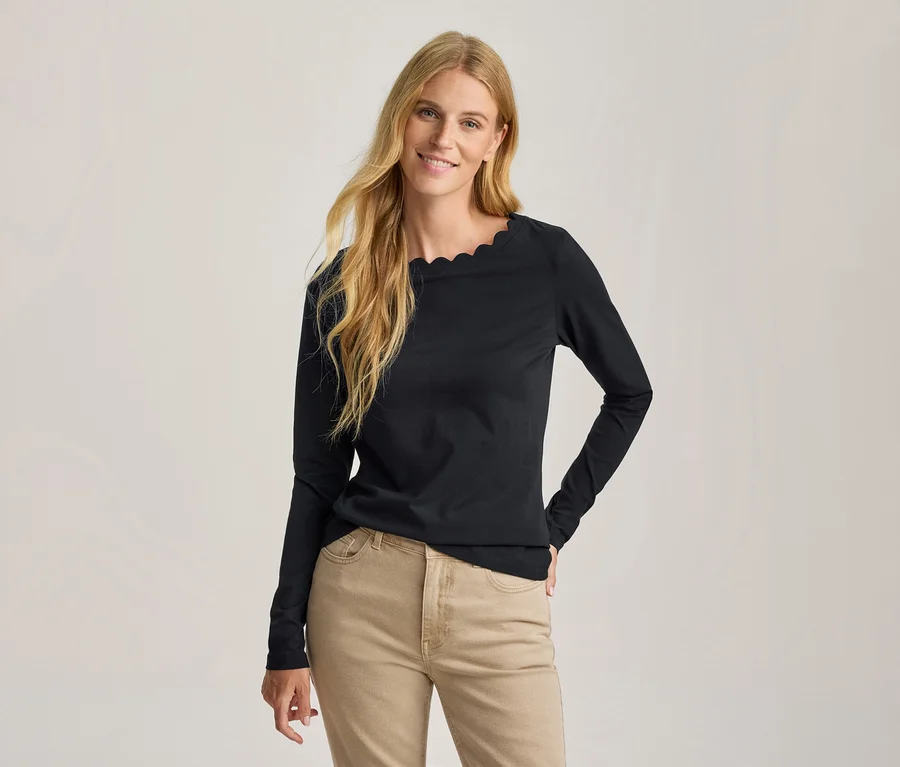 Femme souriante aux longs cheveux blonds portant un haut noir à manches longues avec un décolleté festonné et un pantalon marron clair, une main sur la hanche.