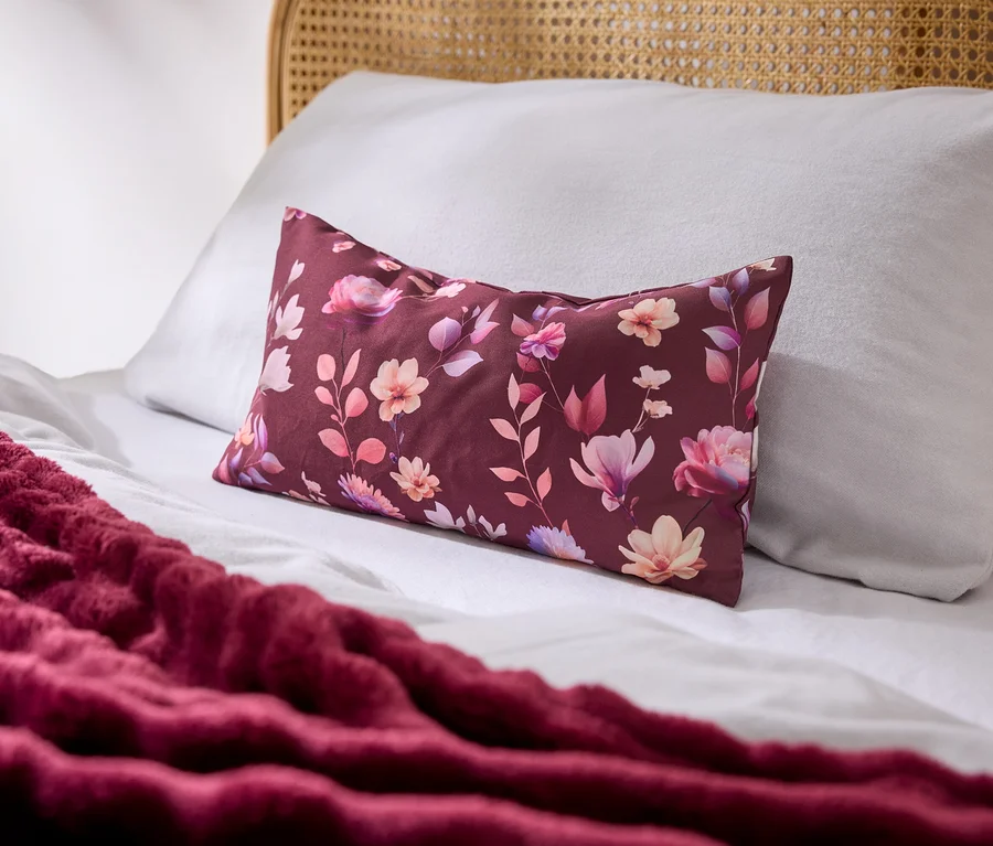 Un lit avec des draps blancs, un oreiller blanc et un oreiller rectangulaire bordeaux à motifs floraux. Une couverture bordeaux texturée est drapée sur le lit, avec une tête de lit en rotin visible.