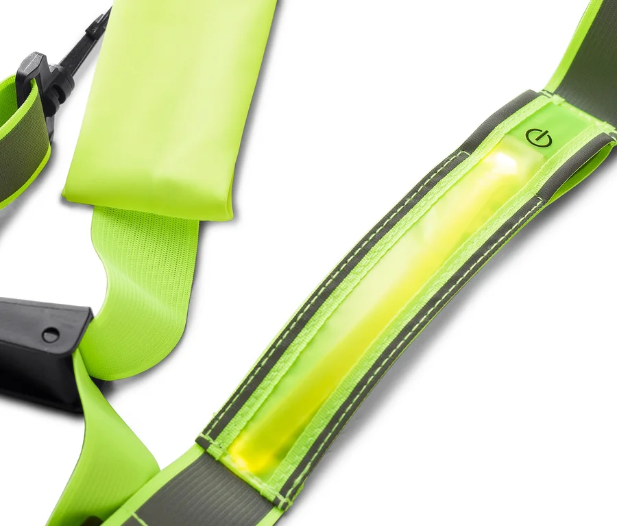 Gros plan d'une sangle de sécurité réfléchissante jaune fluo avec une bande lumineuse et une icône de bouton d'alimentation. La sangle a des bords réfléchissants gris et des coutures noires, avec une boucle noire visible en arrière-plan.