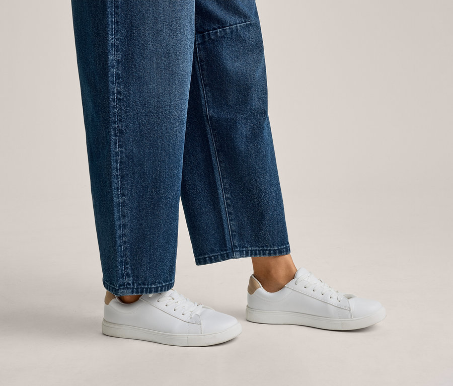 Personne portant un jean bleu et des baskets blanches avec des accents beiges au talon.
