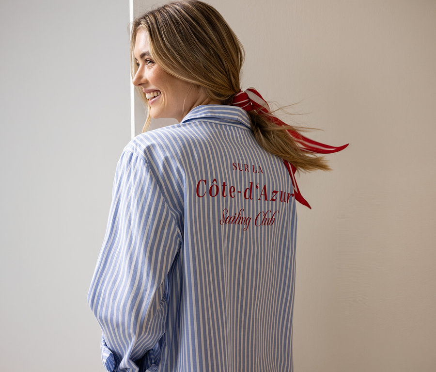 Une femme portant un chemisier Street One rayé bleu et blanc, avec la broderie rouge "SUR LA Côte-d'Azur Sailing Club" dans le dos, sourit en regardant par-dessus son épaule. Un ruban rayé rouge et blanc est noué dans ses cheveux.