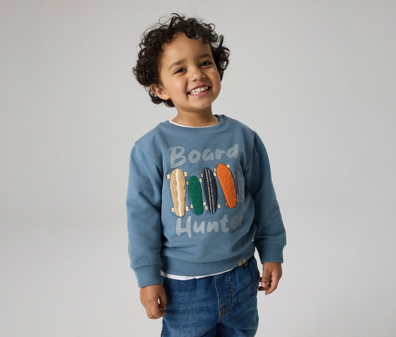 Petit garçon souriant aux cheveux bouclés, portant un pull bleu avec l'inscription "Board Hunter" et des motifs de skateboards, ainsi qu'un jean.