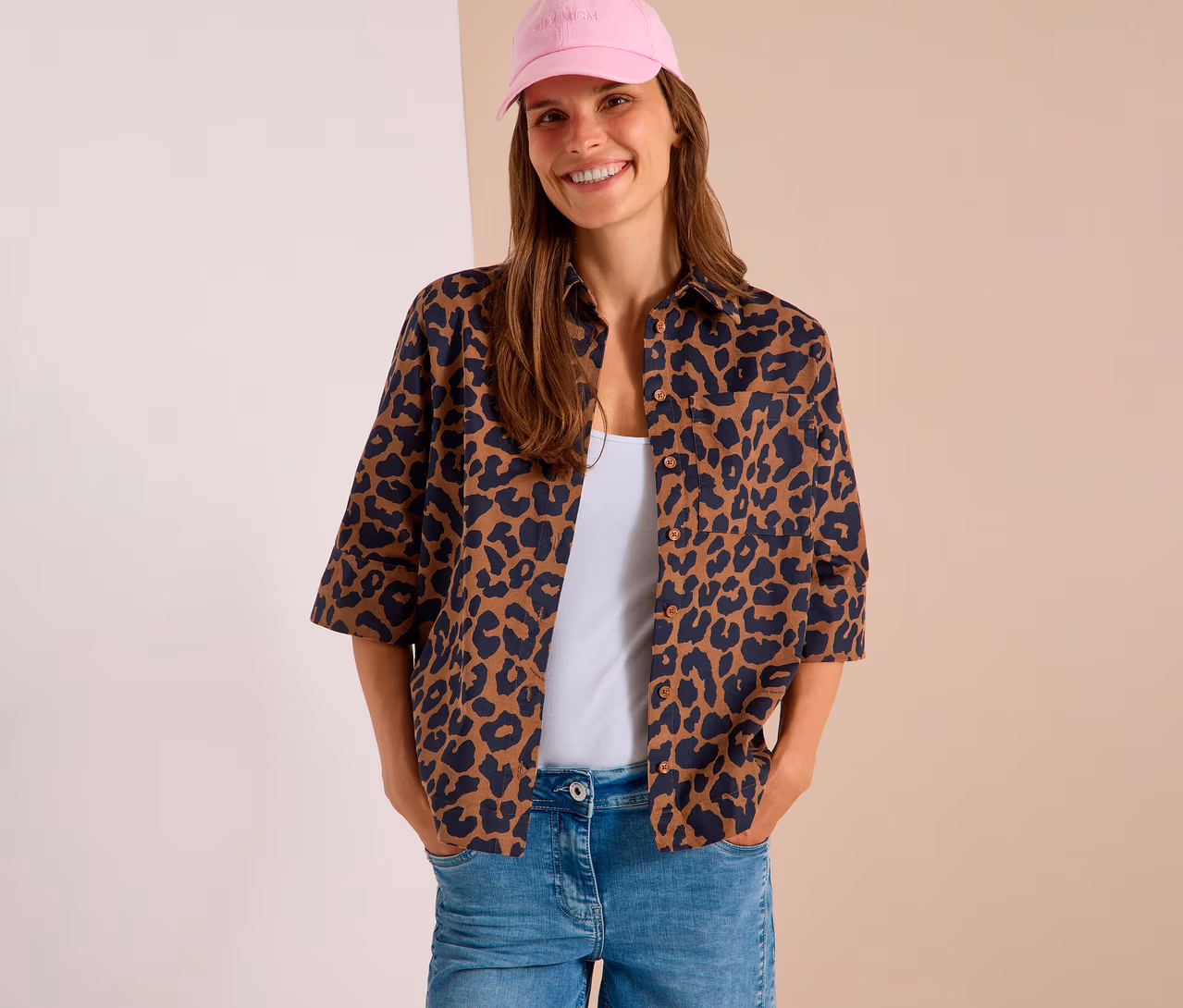 Femme souriante portant une casquette de baseball rose, un chemisier CECIL ouvert à imprimé léopard sur un débardeur blanc et un jean bleu clair, mains dans les poches.