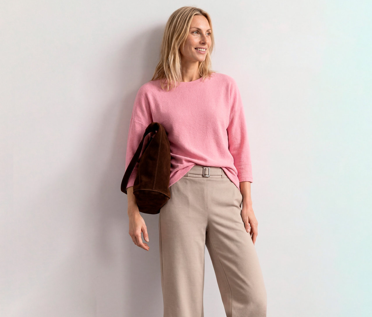 Femme blonde souriante portant un T-shirt à manches 3/4 Street One rose, un pantalon beige et un sac marron, appuyée contre un mur blanc.