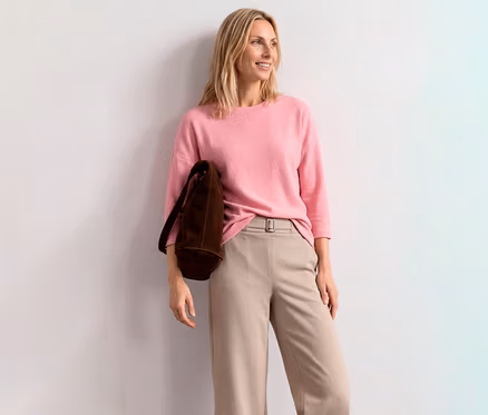 Femme blonde souriante portant un T-shirt à manches 3/4 Street One rose, un pantalon beige et un sac marron, appuyée contre un mur blanc.