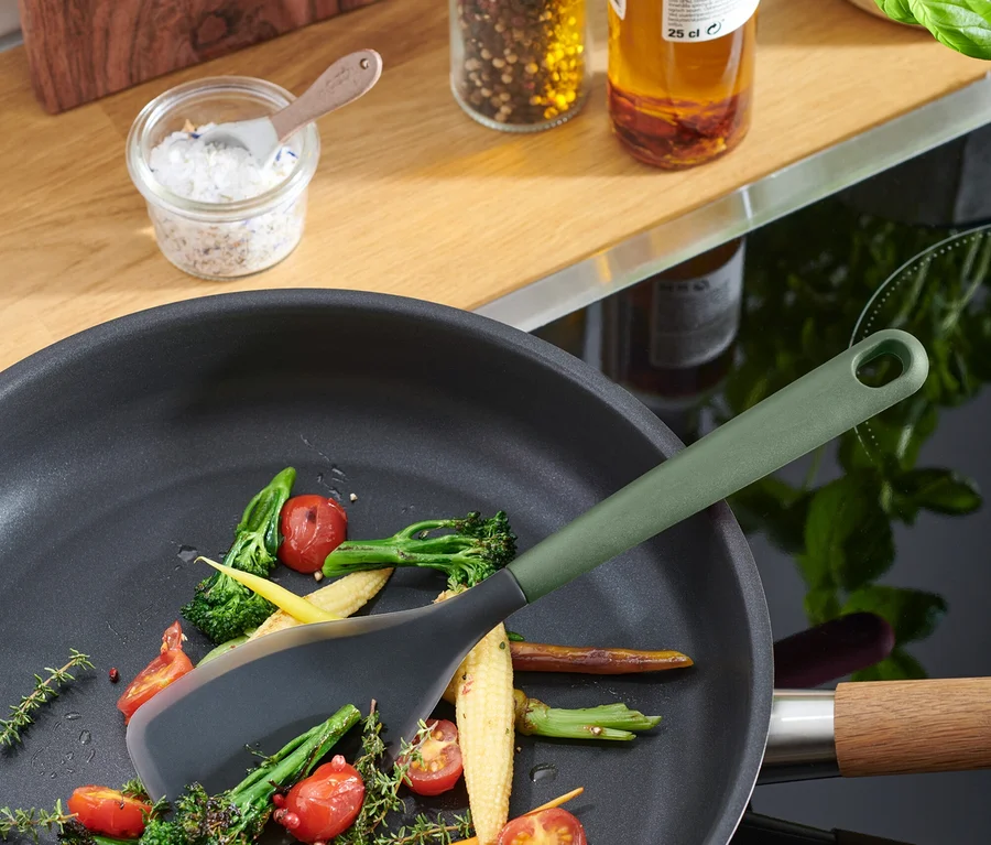 Des légumes sont sautés dans une poêle à frire noire avec une spatule grise à manche vert olive.