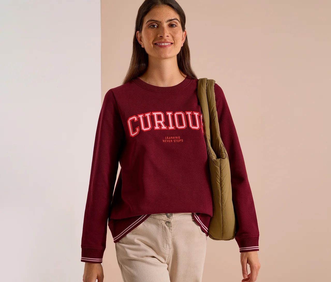 Femme souriante portant un Sweatshirt CECIL bordeaux avec l'inscription "CURIOUS LEARNING NEVER STOPS" et un pantalon beige, un sac vert sur l'épaule.