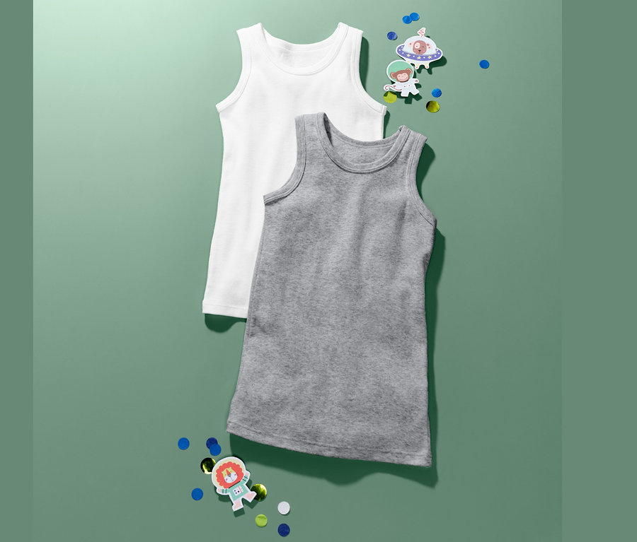 Deux maillots de corps garçon, un blanc et un gris, sont posés sur un fond vert avec des confettis et des autocollants.