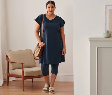 Une femme debout porte une robe en jersey bleu marine avec broderie ajourée, un legging court, des sandales à plateforme en cuir et un sac.