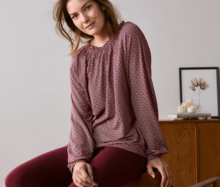 Une femme est assise sur une chaise et porte une blouse imprimée rouge foncé.