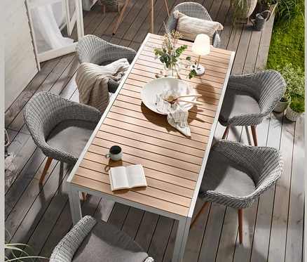 Sur une terrasse de jardin, une table à étirer « Liska » en aluminium est entourée de chaises à coque avec tressage en polyrotin. À l'arrière-plan, un lampadaire à LED sur batterie aspect bois et des pots de fleurs beiges et gris clair avec pieds.