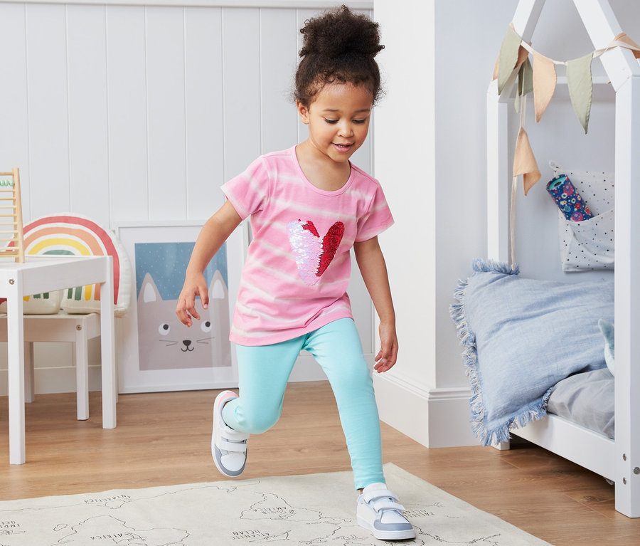Une fillette court sur un tapis dans une chambre d'enfant. Elle porte un t-shirt en coton bio avec un cœur à paillettes.