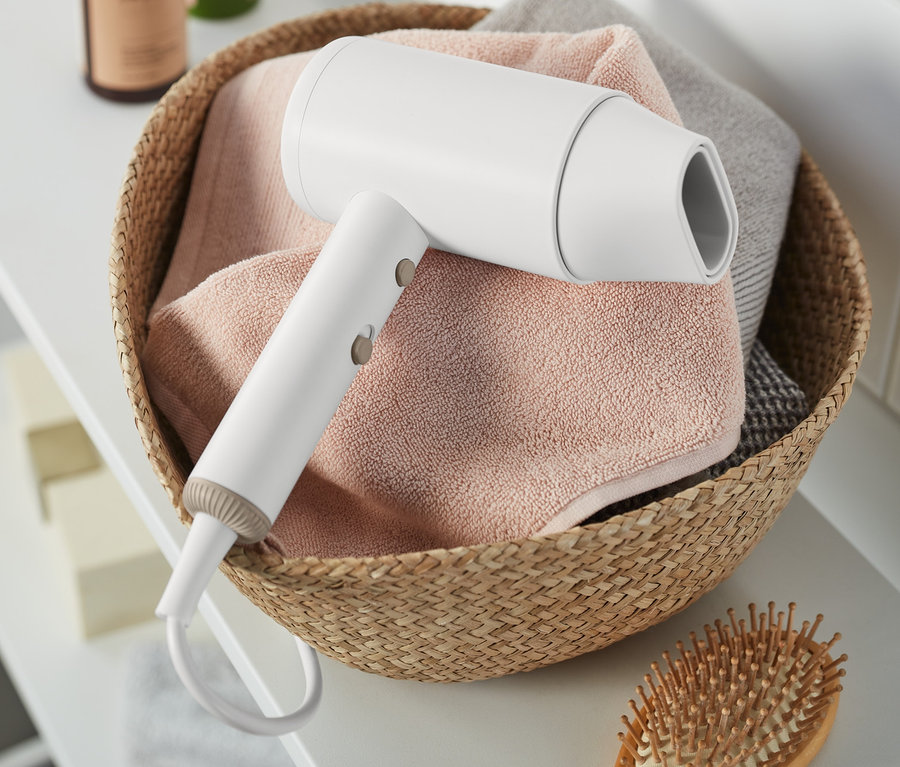 Sèche-cheveux blanc dans un panier en osier avec des serviettes et une brosse à cheveux en bois sur une étagère blanche.