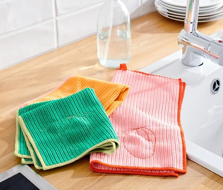 Sur un comptoir de cuisine reposent des 3 chiffons de nettoyage universels verts, oranges et roses à côté d'un évier et de bouteilles d'huile.