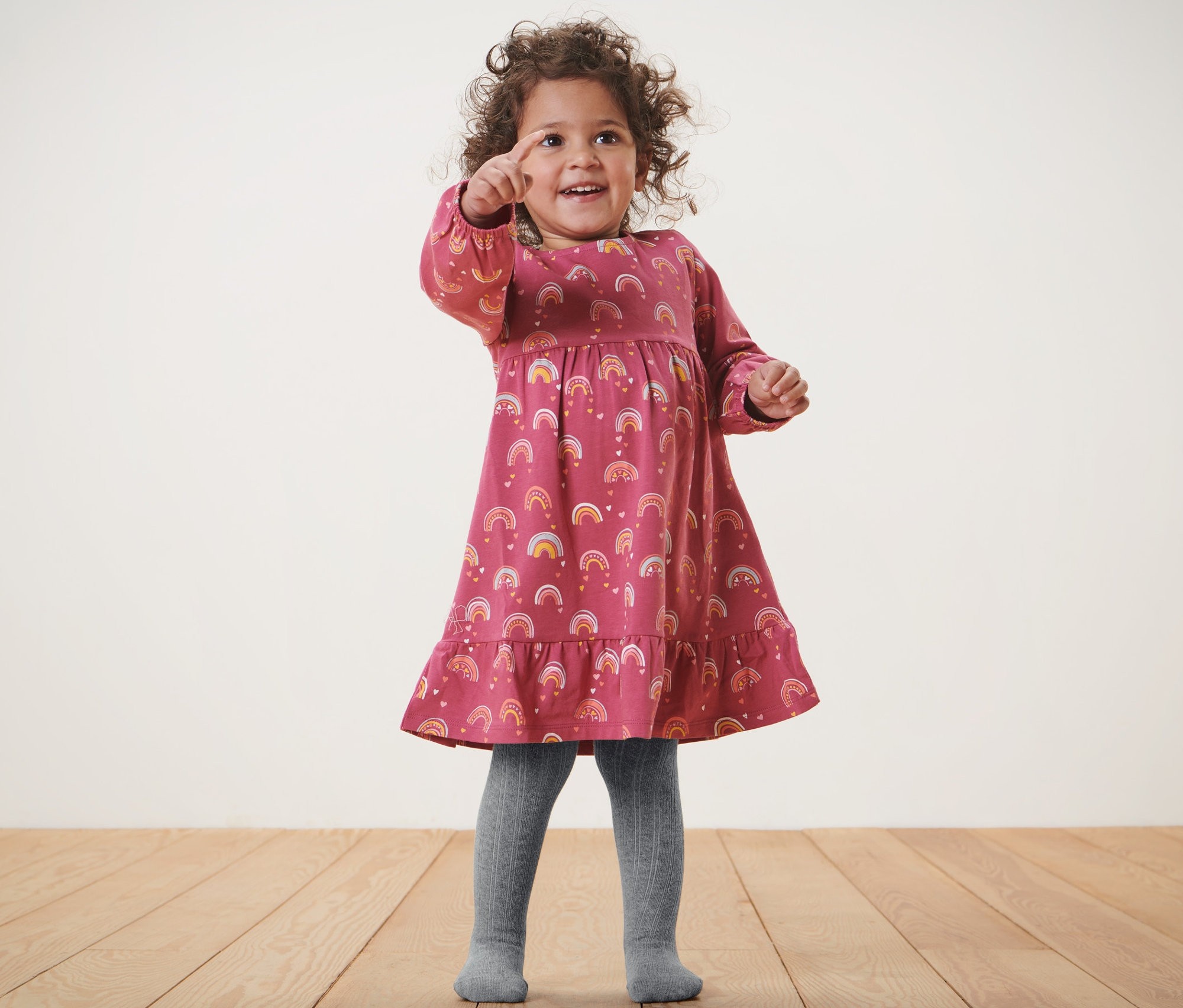 Fillette portant une robe à imprimé arc-en-ciel et 2 paires de collants gris pour bébé pointant du doigt.