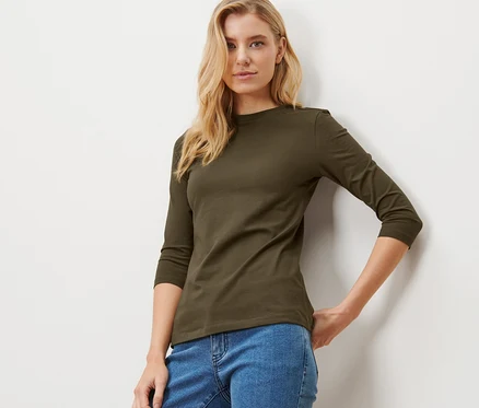 Une femme blonde pose, portant un t-shirt kaki à manches 3/4.
