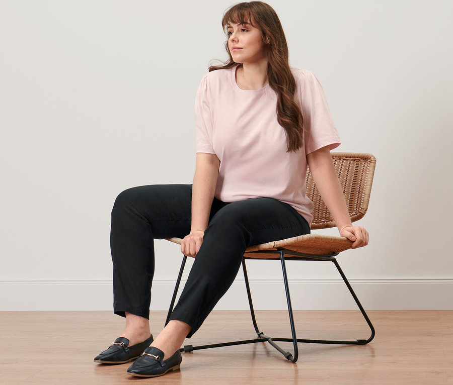 Une femme est assise sur une chaise et porte une chemise rose avec des fronces et un pantalon noir prêt à porter en bengaline.