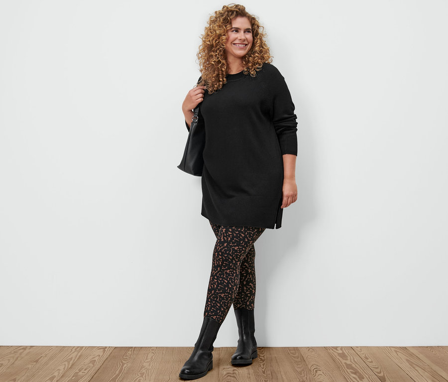 Femme posant dans un long pull en maille noir, un legging imprimé, des bottines en cuir et avec un sac à bandoulière.