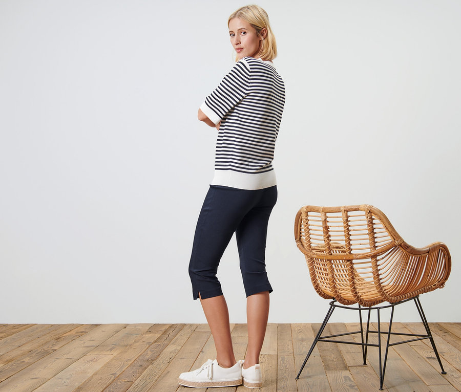Femme posant en pantalon 3/4 stretch bleu foncé et haut rayé à côté d'une chaise en osier.