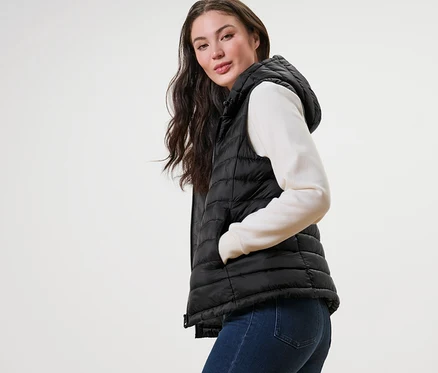 Femme posant en gilet sans manches matelassé à capuche, pull et jean.